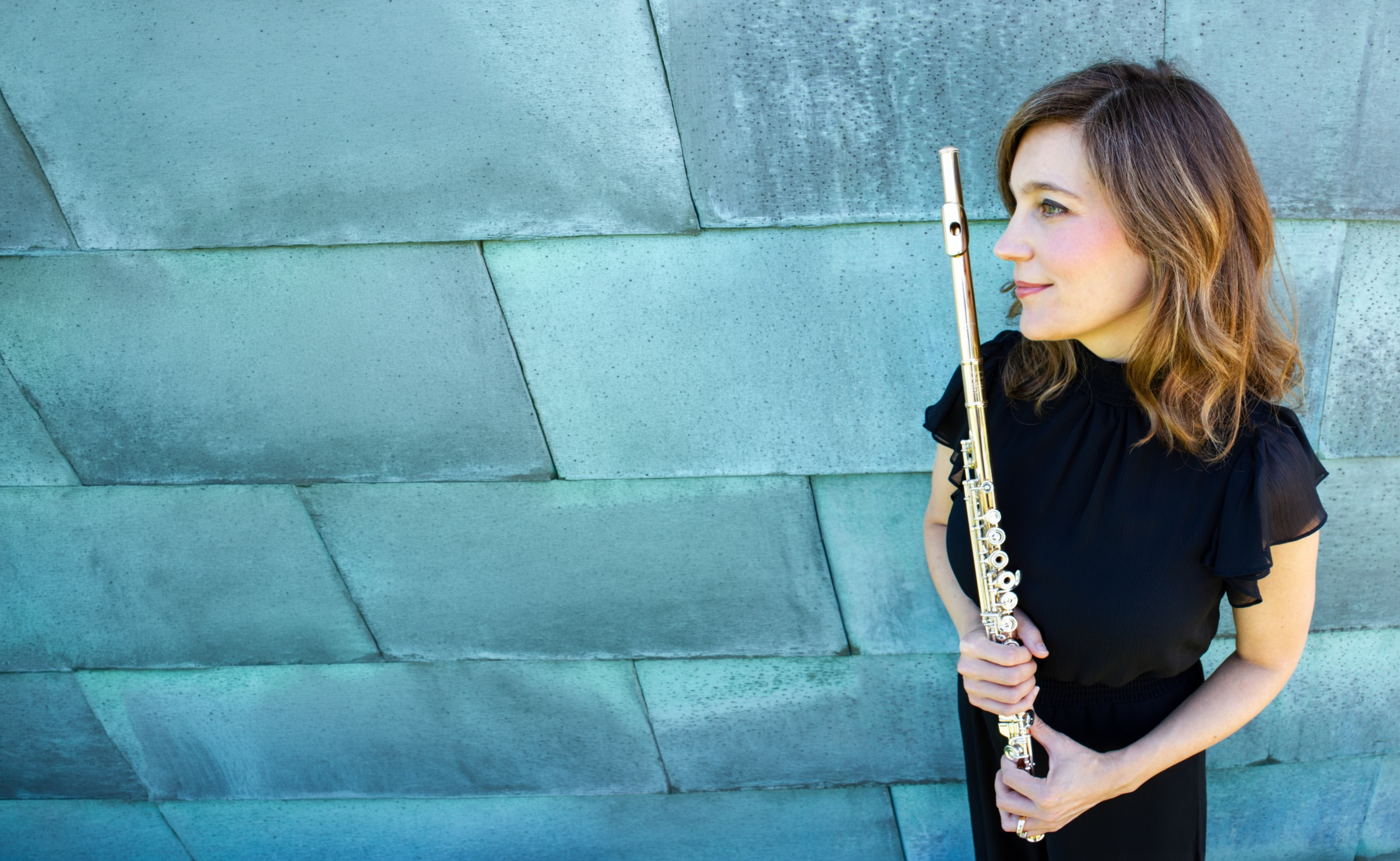 Abigail Walsh holding her flute against a blue-green concrete wall.